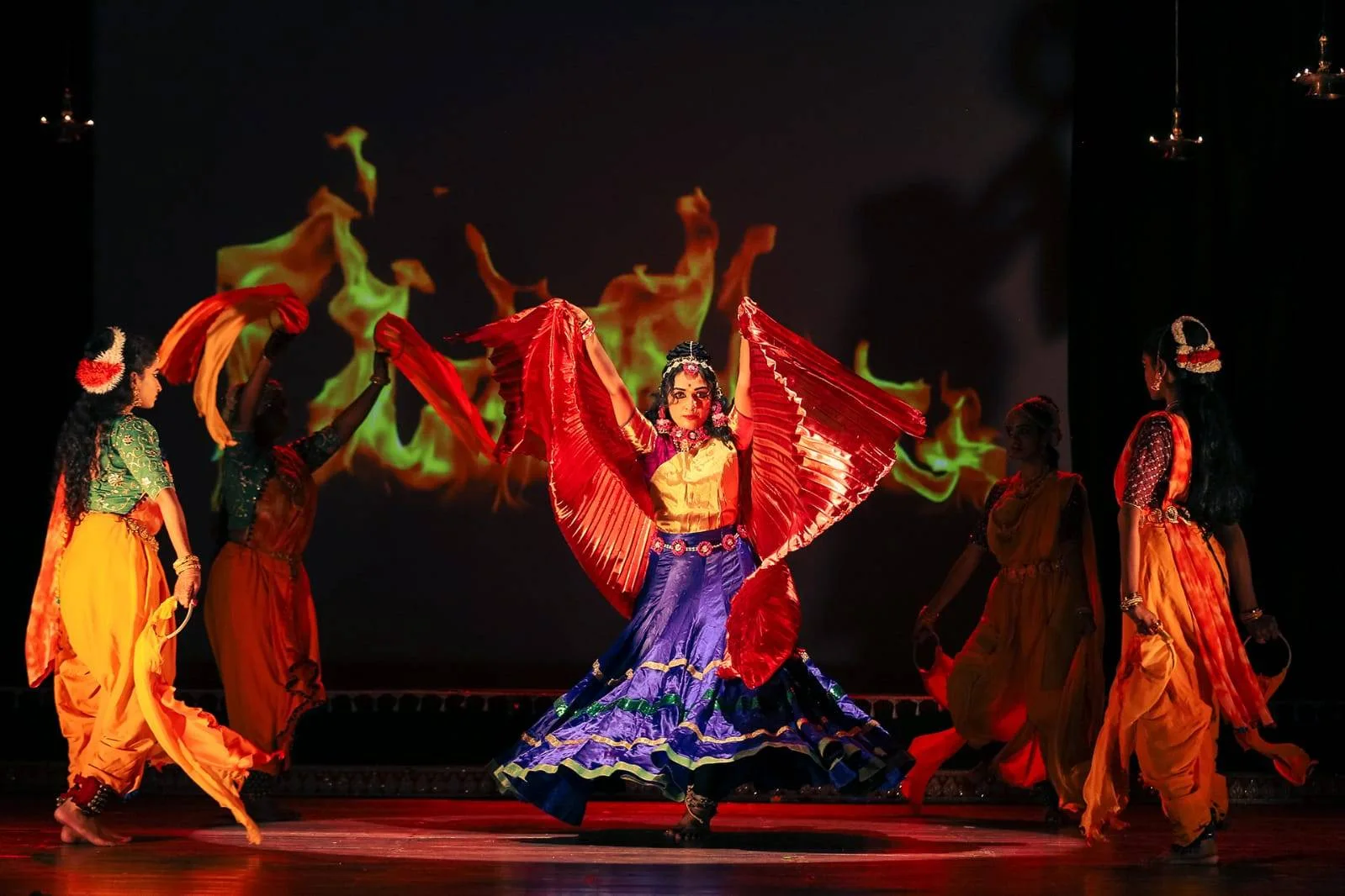 Lead dancer in a red silk veil and violet skirt, surrounded by performers before a projected backdrop of flames