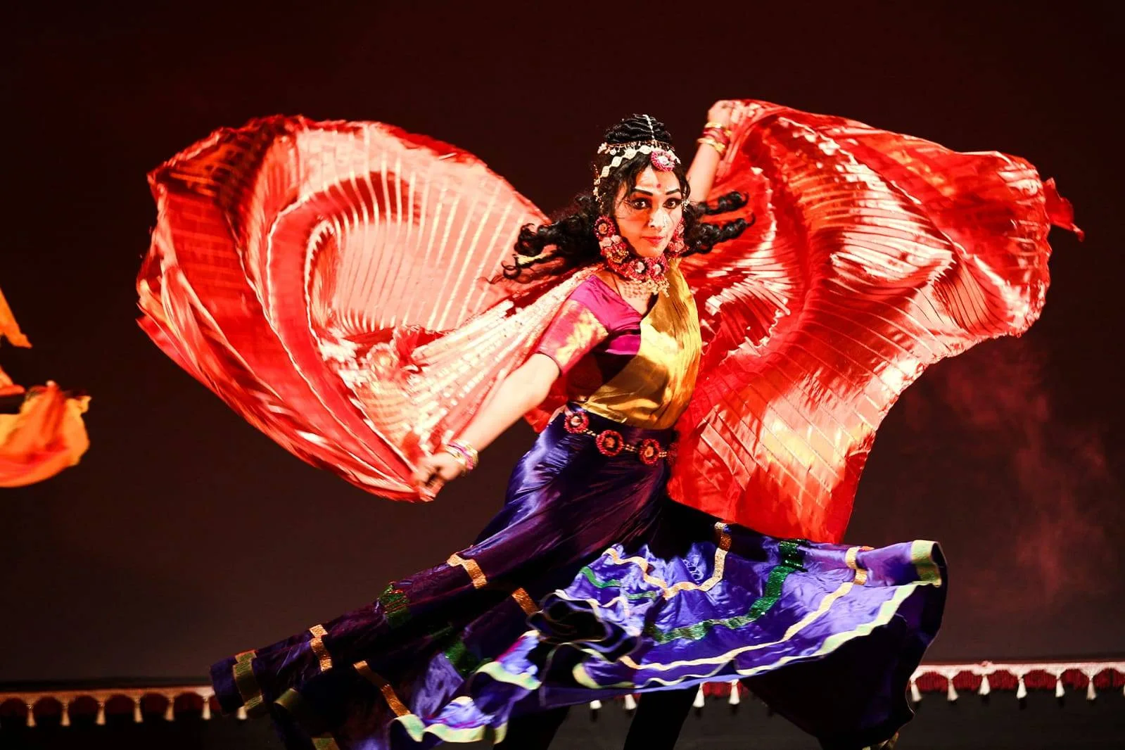 A dancer mid-spin, sweeping pleated red silk wings above a swirling violet skirt
